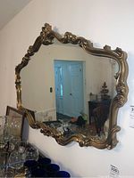 Full view of the large wall mirror showing its ornate gold-colored frame with intricate scrollwork and floral motifs. The mirror reflects part of the room including doorway and furniture.
