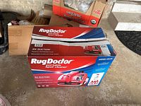 Original packaging box for Rug Doctor portable spot cleaner showing front and side branding, with graphics of the unit and cleaning bottles.