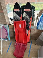 Two Mogul Monster snowboards with black and red mountain design and two plastic sleds, one red and one pink, stacked together.