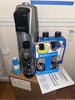 Full view of SodaStream machine with two carbonating bottles in packaging, one loose bottle, CO2 cylinder, instruction manual, all sitting on original box.