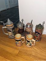 Photo showing set of six German-style drinking vessels: four lidded steins and two unlidded mugs, all with colorful relief decoration on ceramic bodies and metal lids on steins.