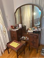 Full view of vintage wooden vanity with tri-fold mirror and needlepoint bench in nearby placement, showing various items on top and some drawer and cabinet details.