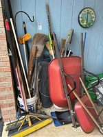 Photo of all yard tools and two wheelbarrows arranged against wall showing all items together with various brushes, brooms, spades, squeegee, and yard hook visible.