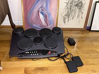Full view of Yamaha DD50 digital percussion MIDI pad drum machine with seven drum pads and connected foot pedals, placed on wooden floor with framed artwork in background.