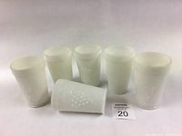 Six white milk glass tumblers standing upright with one tumbler laying down showing the embossed grape and vine motif.