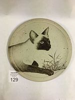 Photo shows round ceramic platter with detailed Siamese cat artwork lying near grass. Speckled texture and matte glaze visible.
