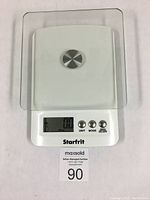 Top view of the white and glass digital kitchen scale powered on showing 0.0 reading with buttons and brand visible.