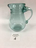 Side view of the translucent light green blown glass jug showing the rounded body, flared rim, rings near the neck, and handle with tear-drop detail.