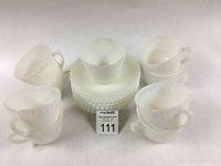 Overview photo showing 9 ribbed translucent tea/coffee cups arranged around a stack of 11 ribbed saucers with petal edges on a white surface.