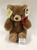 Front view of plush teddy bear with original tags and red neck ribbon, showing its full body and soft brown fur.