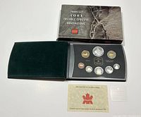 Photo of the opened velvet-like outer case with black insert displaying 8 proof coins, documentation card, and verification hologram sheet. Outer cardboard box positioned behind.