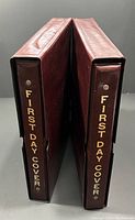 Two maroon First Day Cover albums with gold foil text on spine standing upright within matching slipcases.