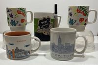 Five Starbucks coffee mugs on a flat surface, with diverse city and floral designs, including Chicago and New York City skylines, and a marker for writing on one mug.