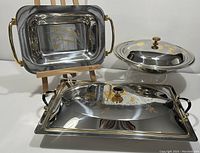 Overview showing all three serving trays: large rectangular tray with gold-tone handles and floral etching, round covered dish with gold floral motif on lid, medium rectangular tray with black handles and gold floral detail.