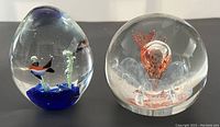 Two Murano glass paperweights side by side on flat surface, showcasing tropical fish design and floral spherical design