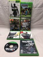Eight Xbox 360 and Xbox One game cases and one disc laid out on a cloth surface, showing covers for Call of Duty Modern Warfare 3, Minecraft, Darksiders, Grand Theft Auto IV, Dying Light, The Witcher 3 Wild Hunt, Call of Duty Black Ops disc, and Call of Duty Ghosts.