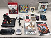 Lot of mixed gaming items including game cases, joystick, PSP, controllers and cartridges arranged on a flat surface.