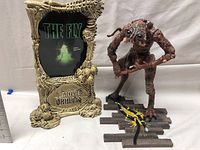 Photo showing the Brundlefly figure standing on a wooden plank base with a detailed backdrop frame displaying 'The Fly' logo and Movie Maniacs branding.