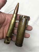 Hand holding both items: a bullet casing bottle opener with brass finish and a pointed tip, and a cylindrical trench lighter with flint striker.