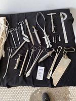Photo showing full assortment of stainless steel medical instruments on black cloth including scissors, scalpel, syringes, forceps, bone saw, and measuring tools alongside canvas wrap.