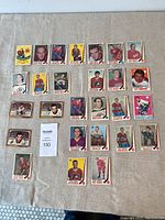 Photo showing 28 assorted vintage hockey cards laid out on a beige fabric surface including players from Flyers, Bruins, Red Wings, Canadiens, Rangers, Kings, Seals, and Canucks.