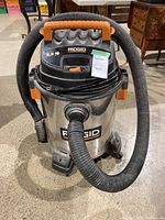 Front view of Rigid shop vac showing 6.5HP and 16-gallon labeling, hose connected at front, and power cord coiled on handle.