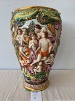 Front view of tall Capodimonte vase showing detailed relief figures of classical adults and cherubs, set in a garden scene with trees and greenery. Multicolored glazes with gold accents visible.