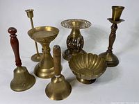 Seven brass decor items including two bells with wooden handles, three differently styled brass candle holders, one scalloped edge brass bowl, and one large decorative brass piece with ornate cutouts.