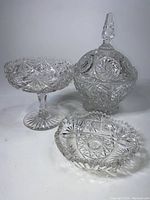 Three crystal pieces displayed on a white surface showing their overall shape and cut patterns: pedestal dish, covered candy dish, and scalloped bowl