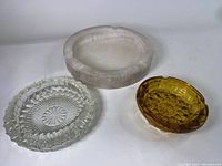 Three ashtrays placed on white surface: white marble, clear glass with pattern, amber glass ashtray