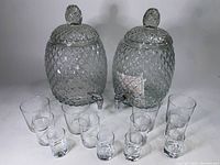Two pineapple-shaped glass drink dispensers with lids and spigots arranged behind a fan of 12 assorted clear drinking glasses.