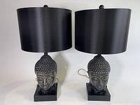 Pair of Safavieh Buddha head table lamps with black cylindrical shades standing side by side on a white background.