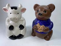Front view showing both cow and bear ceramic cookie jars side by side on white surface.