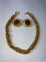 Gold tone chain link necklace with toggle clasp and matching clip-on earrings with circular design and dark center detail laid flat on white background.