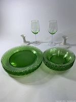 Six green Pyrex plates and six matching green Pyrex bowls displayed alongside two clear wine glasses and two white ceramic candle holders.