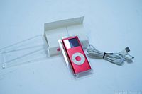 Front view of red Apple iPod nano 2nd Generation in clear plastic case, showing screen and white control wheel, next to white charging cord and original box.