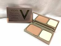 Open compact showing beige powder foundation on one side and white applicator sponge on the other, with mirror inside the lid.