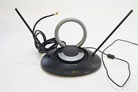 Front view of RCA ANT501 amplified indoor antenna showing black base, circular UHF loop antenna in center, and two VHF dipole antennas on either side with cables attached.