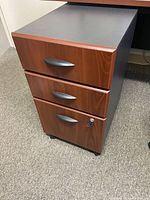 Mobile pedestal with 3 drawers, cherry wood finish with black top, lock on bottom drawer visible, on carpeted floor.