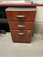 Front view of a three-drawer mobile pedestal with wood finish and silver handles, placed under a desk.