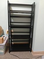 Photo of a black 4-tier freestanding bookshelf placed against a light-colored wall on carpet.