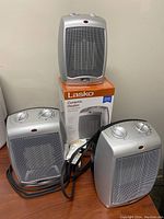 Three silver Lasko ceramic mini heaters, two on table and one on box, with cords and control knobs visible.