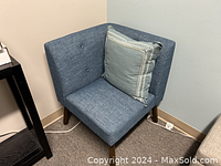 Blue fabric corner chair with a decorative pillow, photographed from an angle showing the seat, backrest, and legs. Pillow excluded from the lot.