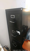 Photo of the black metal file cabinet showing four drawers with metal handles and visible wear, including peeled paint and scratches. Keyhole visible on the top drawer.