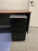 Photo of black 3-drawer metal file cabinet under wooden desk, carpeted office floor visible.