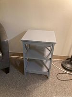 Gray wooden side table with two shelves placed against a beige wall on a carpeted floor. Side table has a flat square top and two shelves with crossbar supports on side panels.