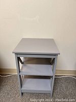 Gray side table with two lower shelves, set against a plain wall with visible power cord at the baseboard.