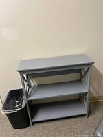 Photo shows a gray wooden side table with three shelves placed against a beige wall next to a small trash bin.