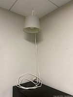 Photo of one white hanging lamp with conical lampshade hanging from ceiling with long white cord coiled on floor against beige walls.