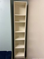 Tall, narrow Ikea Billy bookcase with six shelves, cream/off-white color, standing against walls in a corner.